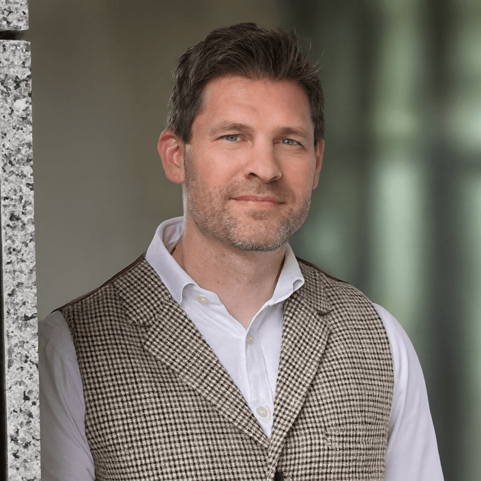 Smiling man in a checkered vest leaning against a stone wall.