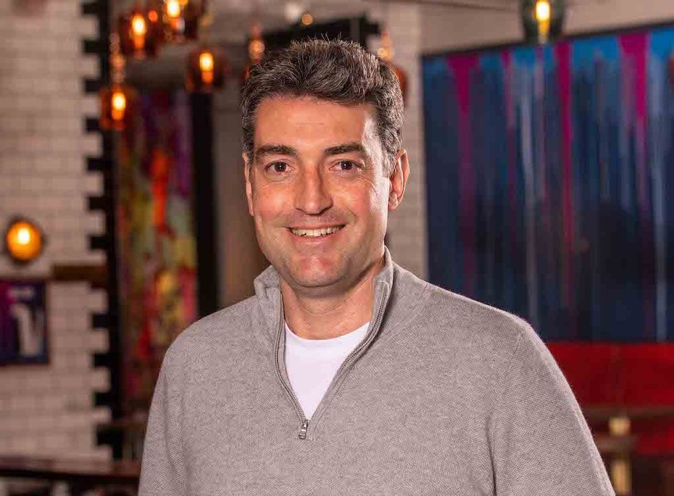 Smiling man in a gray sweater in a warmly lit room with colorful abstract art.