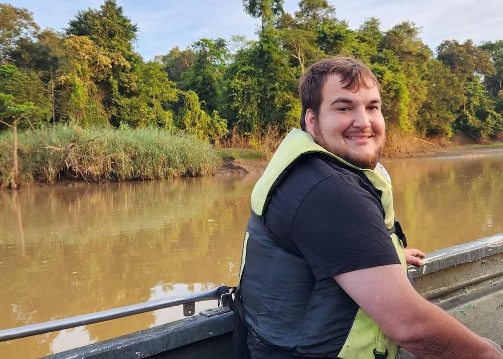 Smiling man in a life vest on a boat, with river and lush trees in the background.