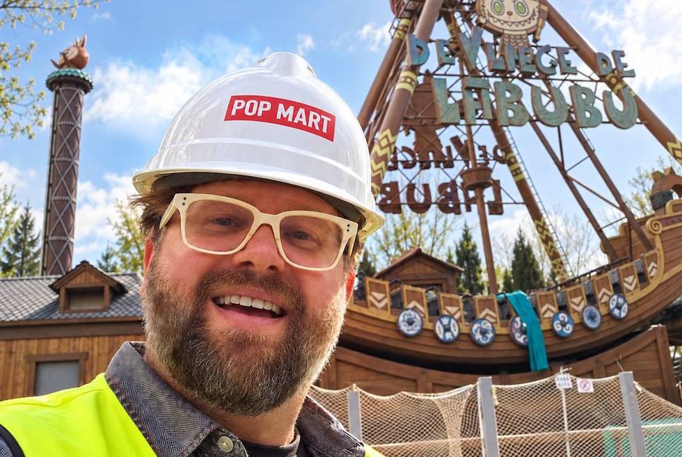 Smiling man in a Pop Mart hard hat and safety vest at a theme park construction site.