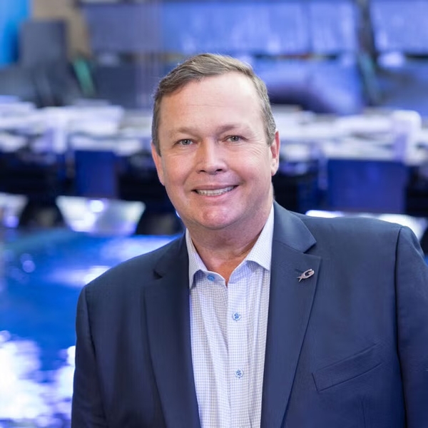 Smiling man in a suit, standing in front of a blurred blue-lit background.