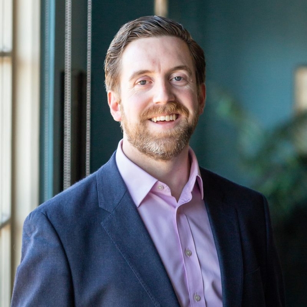Smiling man in a suit with a pink shirt, standing by a window.