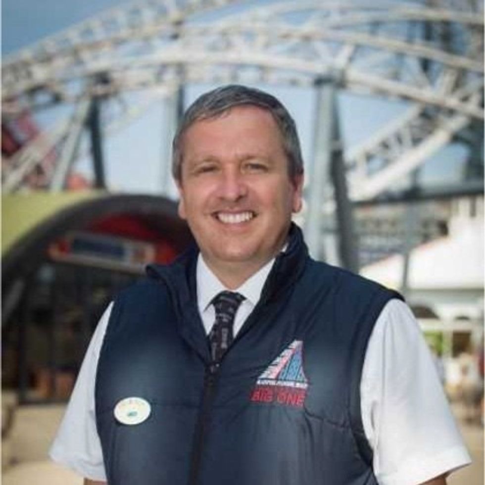 Smiling man in a vest with roller coaster structure in the background.
