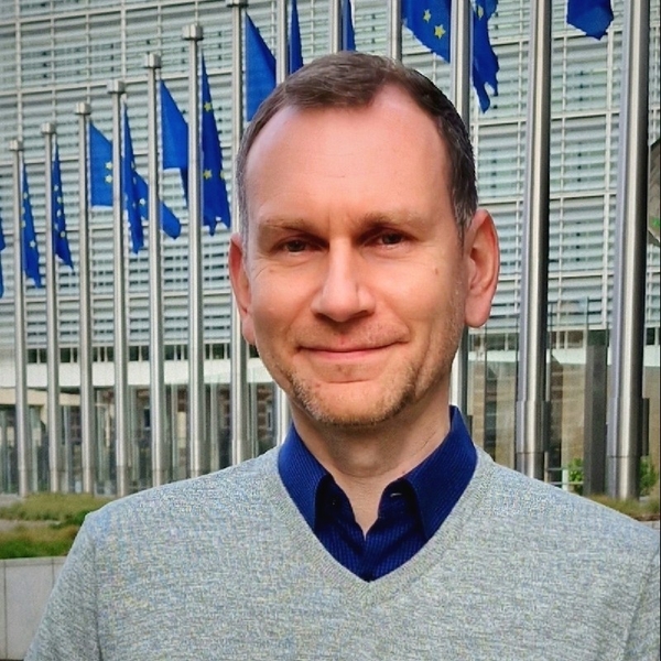 Smiling man in front of EU flags and a modern building.