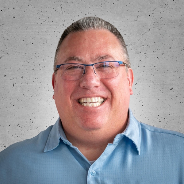 Smiling man in glasses with a blue shirt against a textured gray background.
