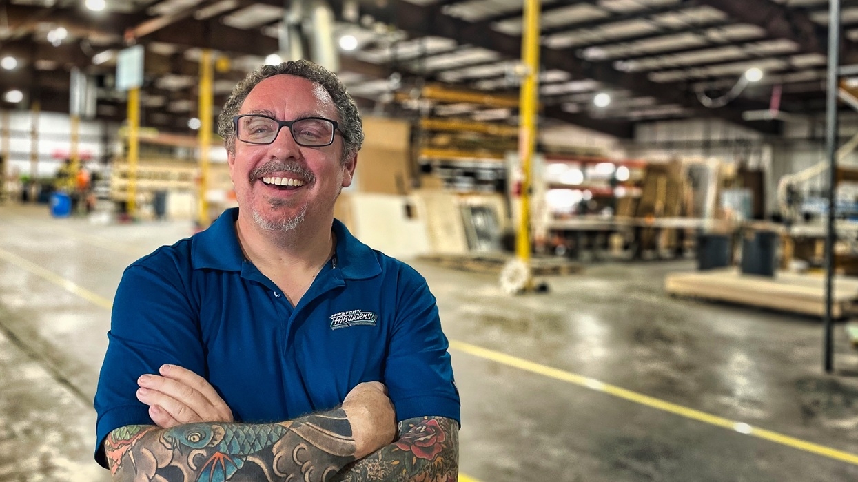 Smiling man named Daniel Krall, with tattooed arms in a warehouse, wearing a blue shirt and glasses.