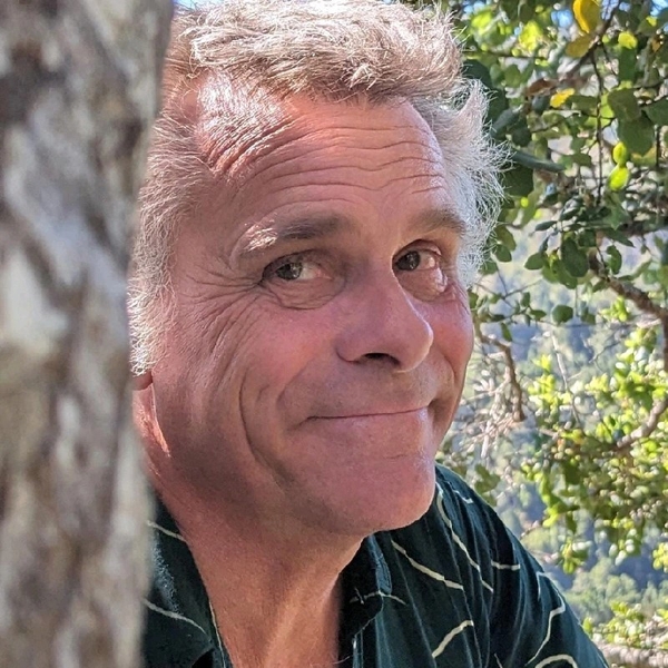 Smiling man peeking around a tree outdoors, with greenery in the background.