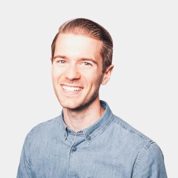 Smiling man wearing a light blue button-up shirt against a plain background.
