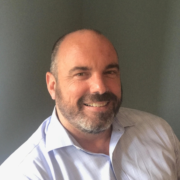 Smiling man with a beard in a light shirt against a plain background.