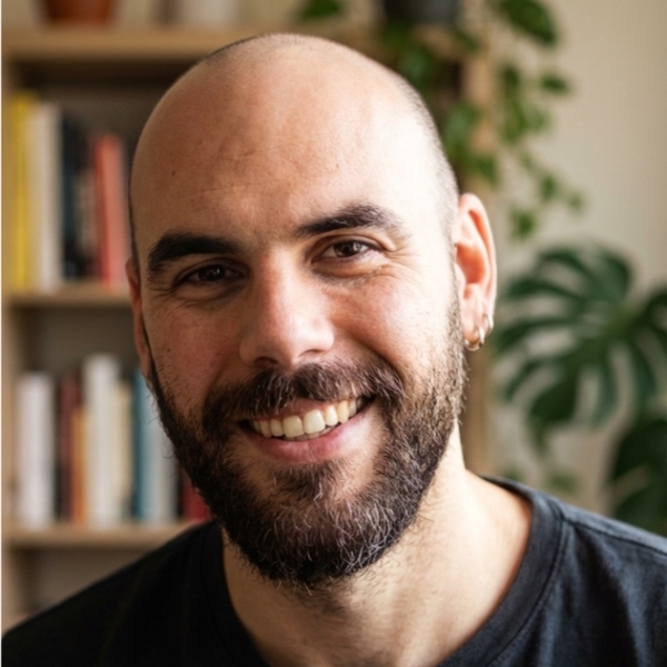 Smiling man with a beard in front of a bookshelf and a leafy plant.