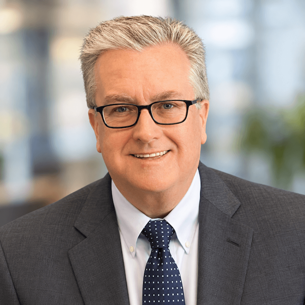 Smiling man with gray hair and glasses in a dark suit and patterned tie.