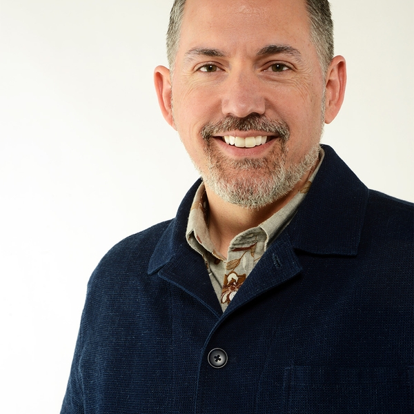 Smiling man with short hair and beard in blue jacket against a light background.