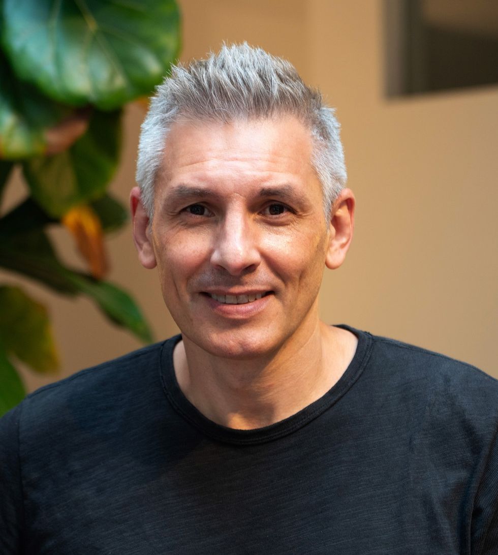Smiling man, Yan Vizinberg, with gray hair in black shirt, standing indoors near a leafy plant.