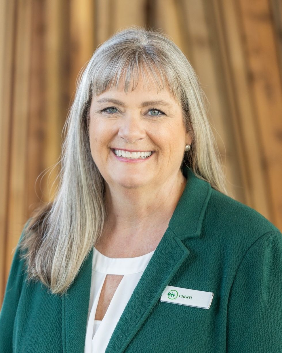 Smiling person, Cheryl Christensen, with long blonde hair, wearing a green jacket and name badge.