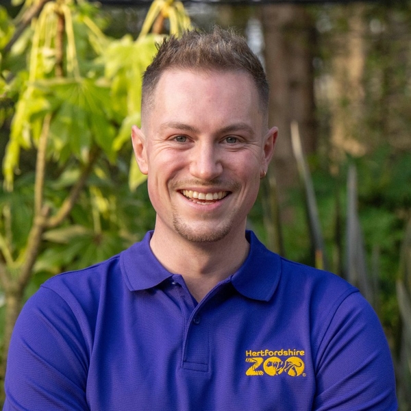 Smiling person in a blue zoo shirt stands outdoors with greenery in the background.