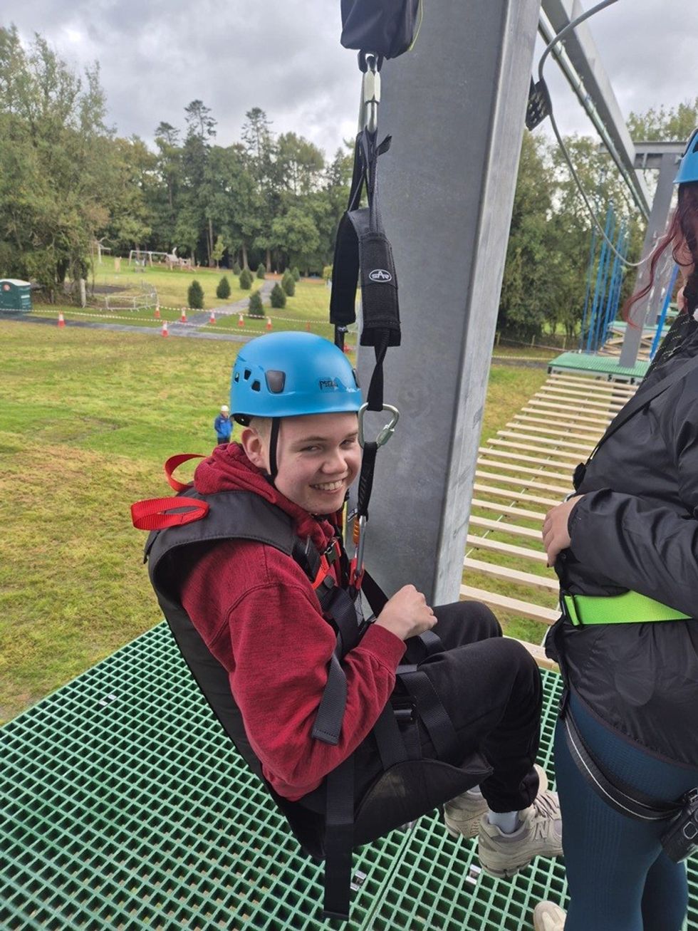 Smiling person in harness, ready for zip-lining, wearing a helmet on a green platform.