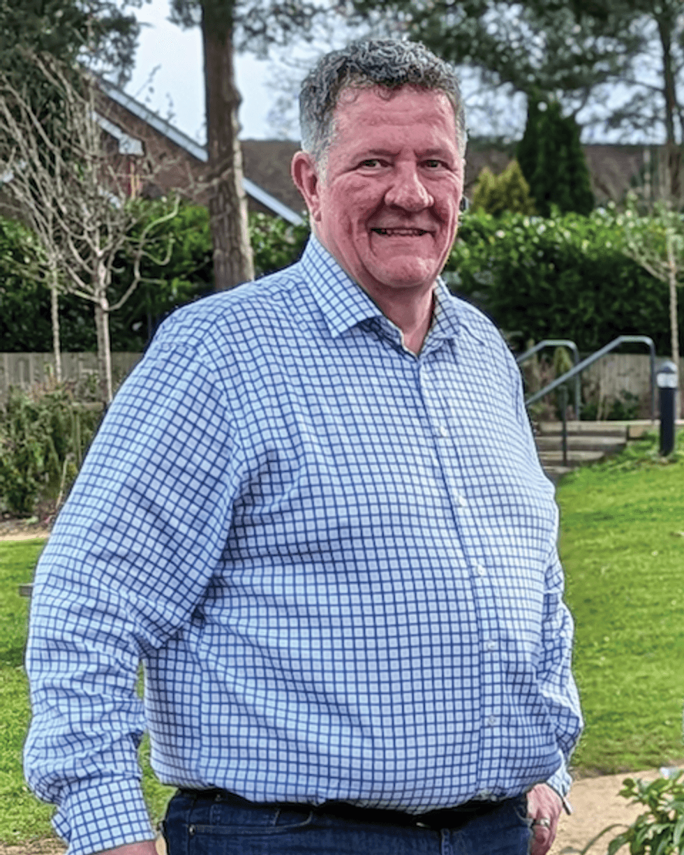 Smiling person named Andy Povey in a checkered shirt standing in a garden with trees and a house in the background.