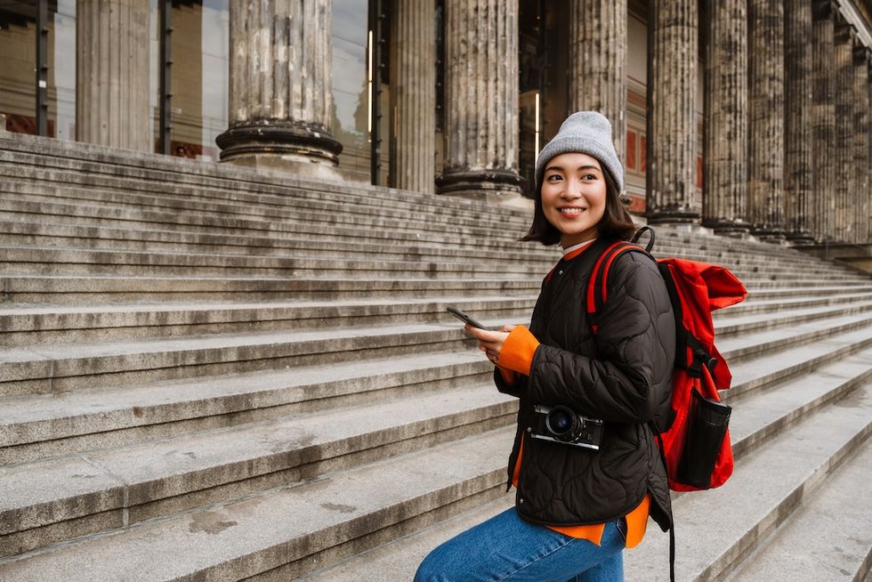 Smiling person with backpack and camera on steps of a grand building.