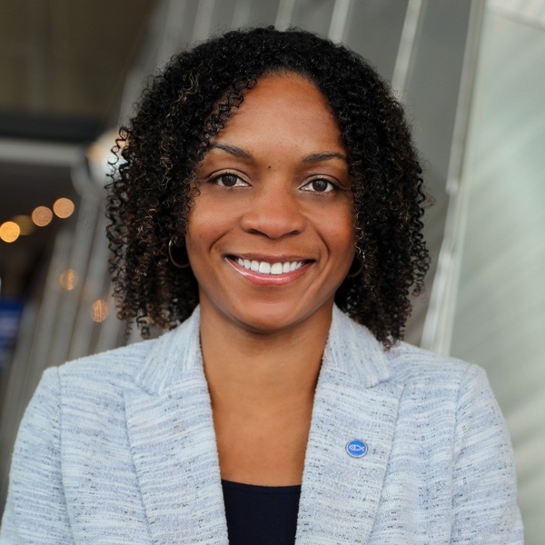 Smiling person with curly hair in a light blazer, standing against a blurred indoor background.