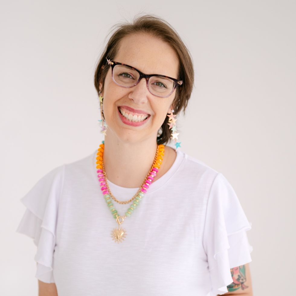 Smiling person with glasses, colorful jewelry, and a white shirt against a plain background.