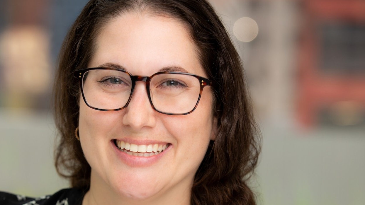 Smiling person with glasses, dark hair, blurred outdoor background.