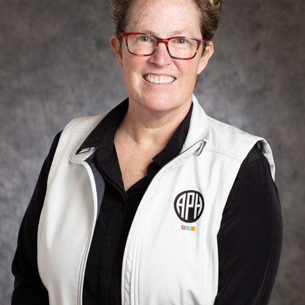 Smiling person with short curly hair, red glasses, in a white APH vest and black shirt.