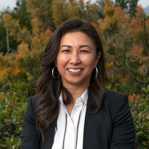 Smiling woman in a black blazer, outdoors with greenery and orange foliage in the background.