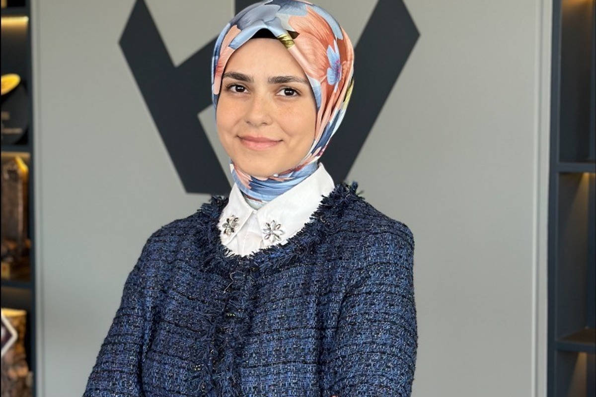 Smiling woman in a floral headscarf and blue jacket, standing with arms crossed.
