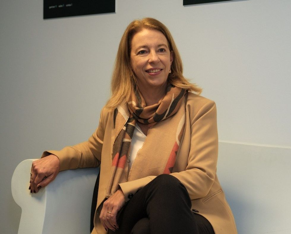 Smiling woman in a tan jacket and scarf sitting on a white chair.