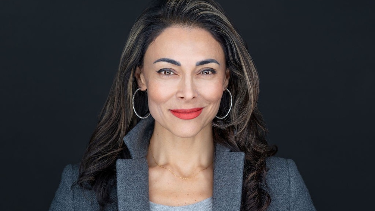 Smiling woman in gray blazer and hoop earrings against a dark background.