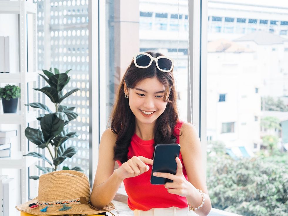 Smiling woman in red uses phone, indoors, with window view and potted plant.