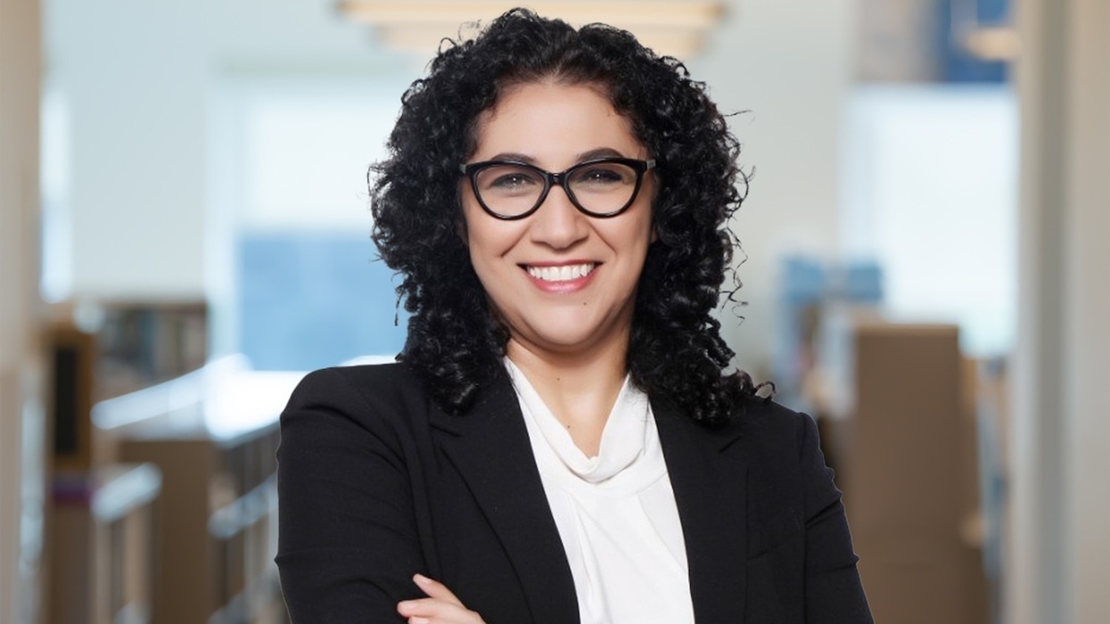 Smiling woman named Irene Eshak, with glasses in a black blazer, standing with arms crossed indoors.