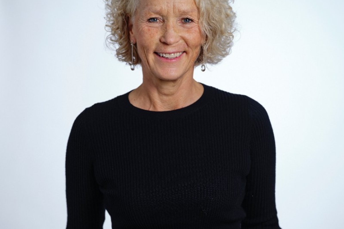 Smiling woman with curly gray hair in a black sweater against a light background.