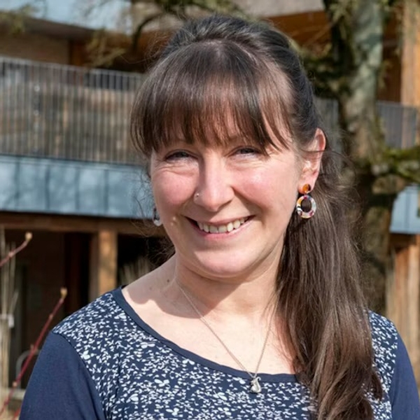 Smiling woman with long hair, wearing patterned top; tree and building in background.