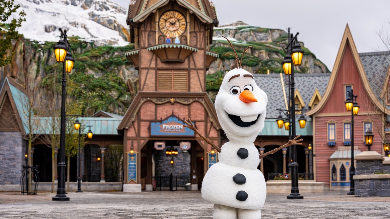 Snowman called Olaf posing at Disney theme park entrance with snowy mountain in the background.