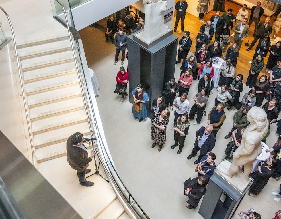 Speaker addressing a crowd in an art gallery by a staircase and sculptures.