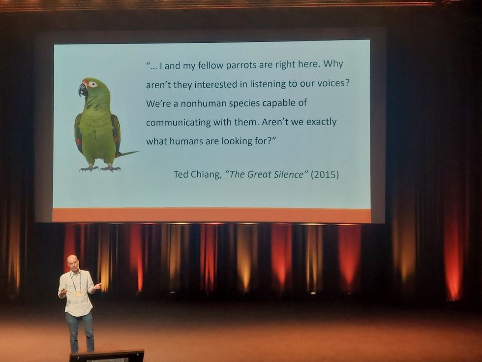 Speaker presenting a slide with a parrot and a quote from Ted Chiang's "The Great Silence".