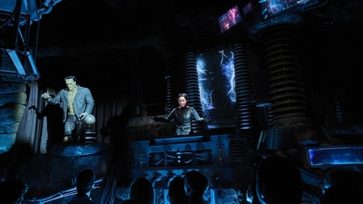 Spooky lab scene from Epic Universe's Monsters Unchained dark ride with Franekstein animatronic, control panel, and glowing electric effects; audience watches.