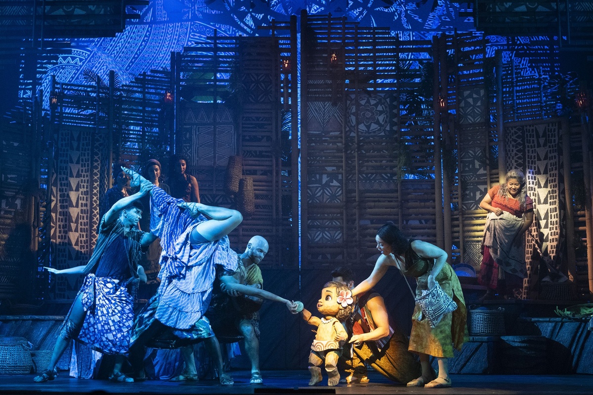 Stage performance with actors interacting with a child puppet, tribal patterns backdrop.