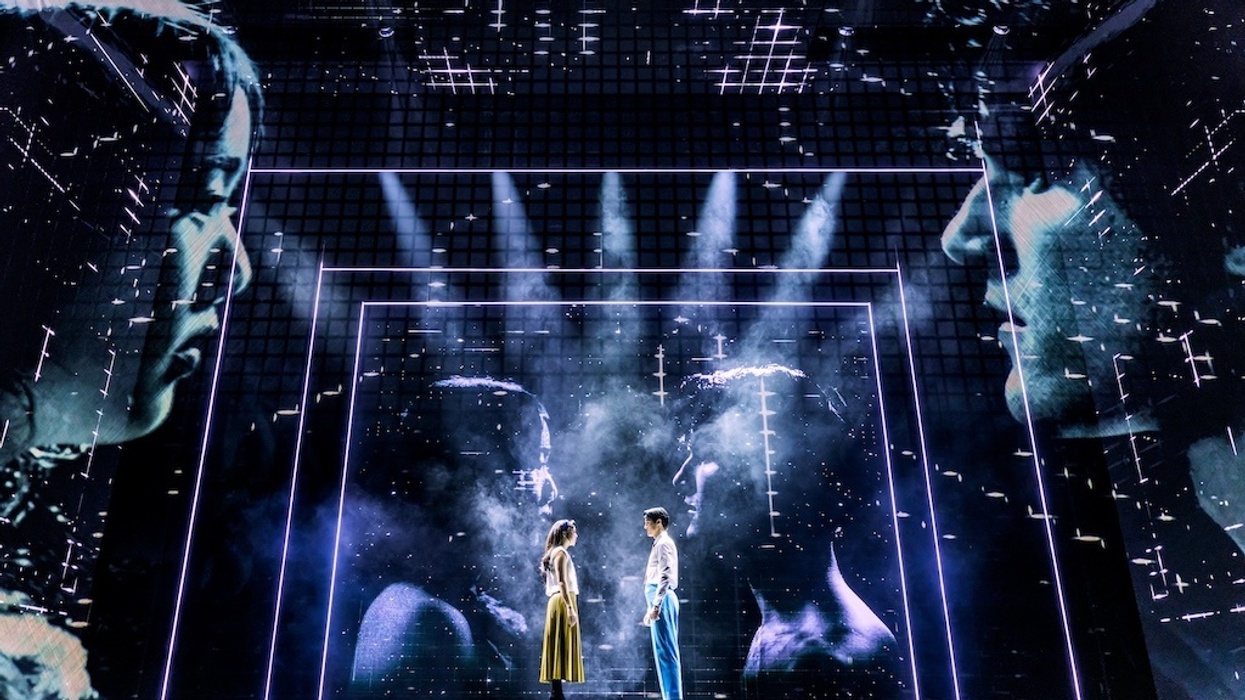 Stage scene with two actors facing each other amid dramatic digital projections.