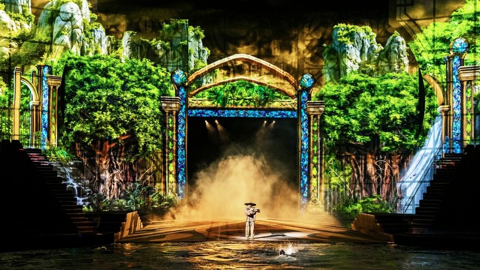 Stage with vibrant forest backdrop and arches in The House of Dancing Water, with mist rising, a performer stands at center.