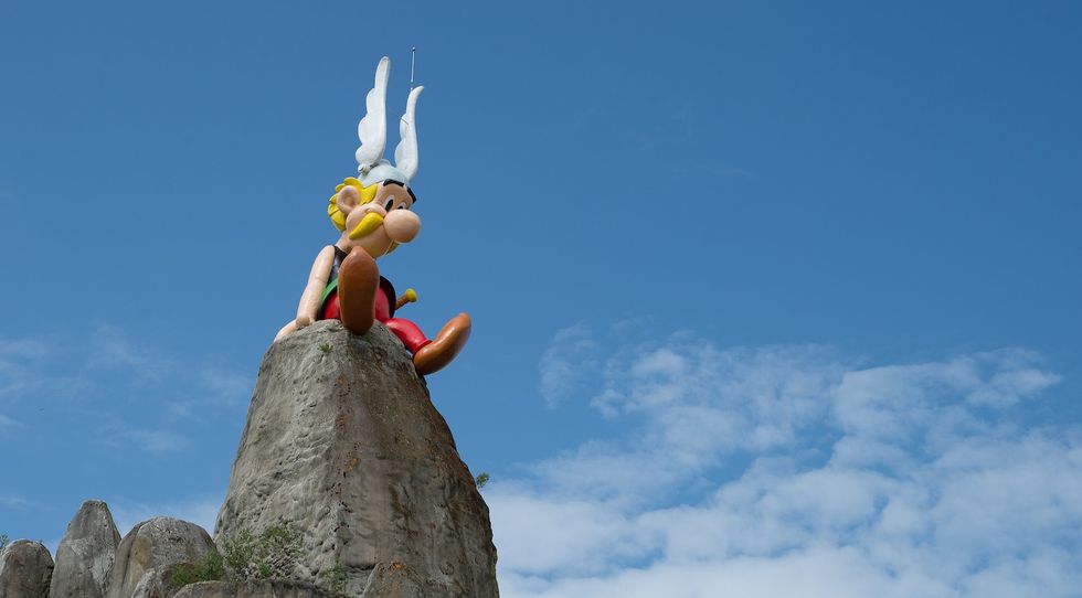 Statue of Ast\u00e9rix cartoon character with helmet atop rock against cloudy blue sky.