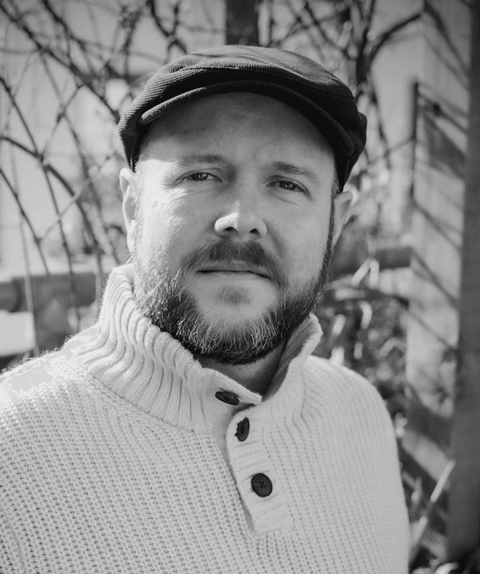 Steve Lance, a man with a beard and cap in a sweater, outdoors, monochrome.