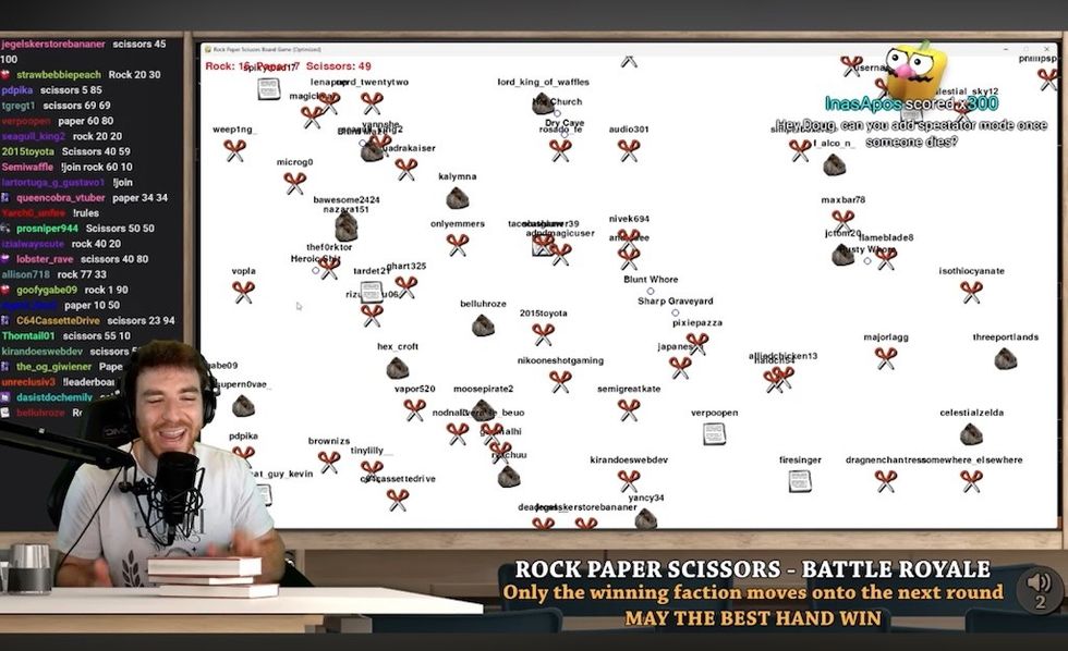 Streamer commentates a Rock Paper Scissors Battle Royale displayed on a large screen.