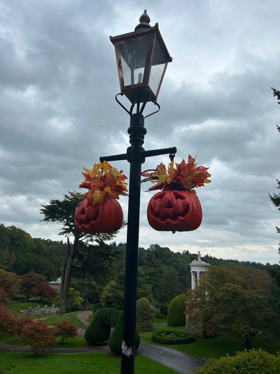 Streetlamp with two hanging jack-o'-lanterns amidst a lush park and cloudy sky.