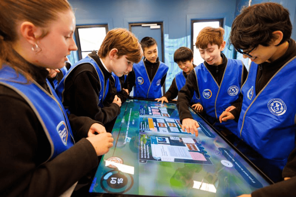 Students interact with a touchscreen table, wearing blue vests.