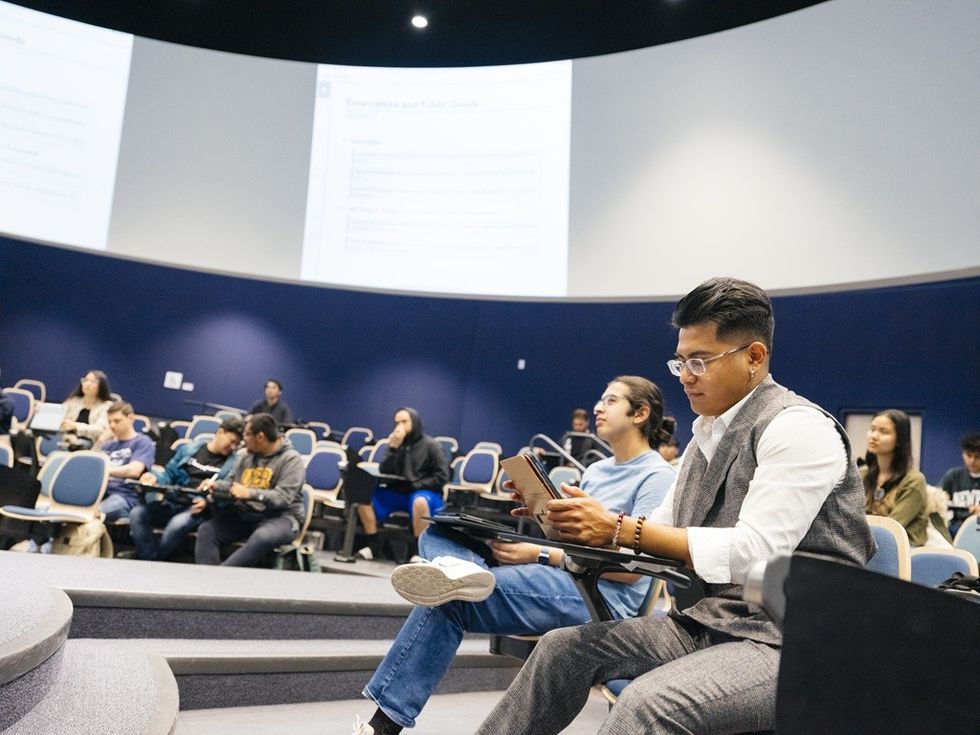 Students sitting in a large lecture hall, some taking notes and others listening attentively.