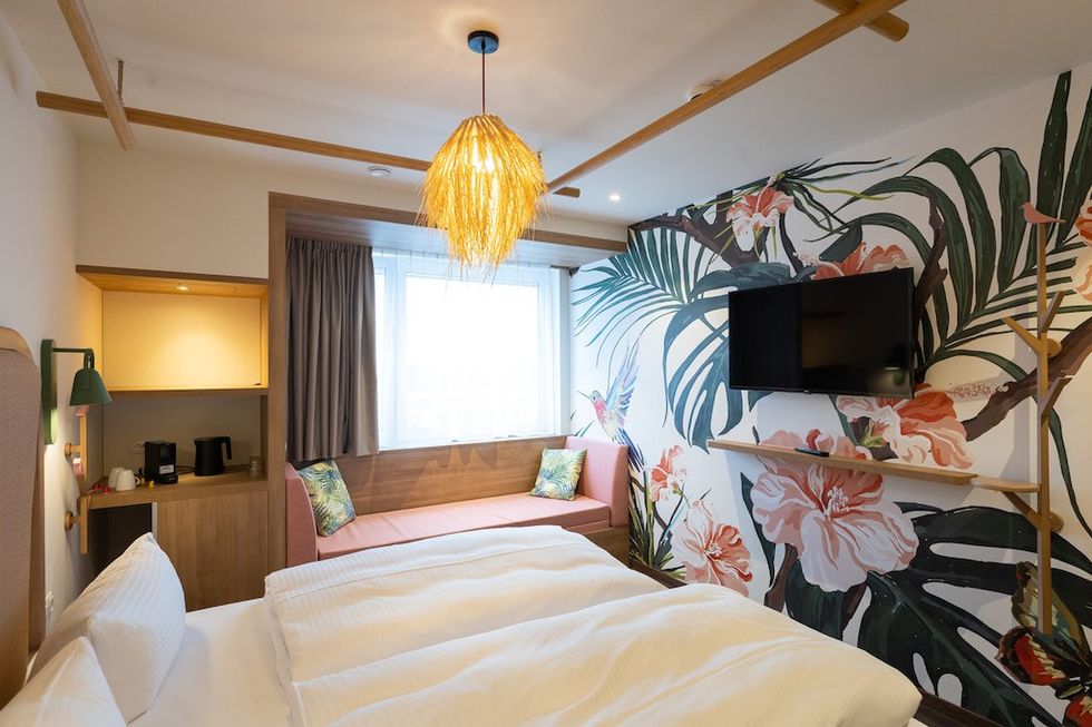 Stylish hotel room with floral mural, bed, sofa, wall-mounted TV, and hanging light fixture.