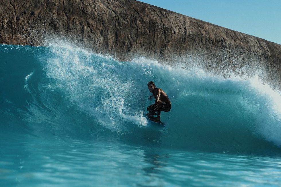Surfer at Adrena carving through a turquoise wave near a rocky barrier under a clear blue sky.