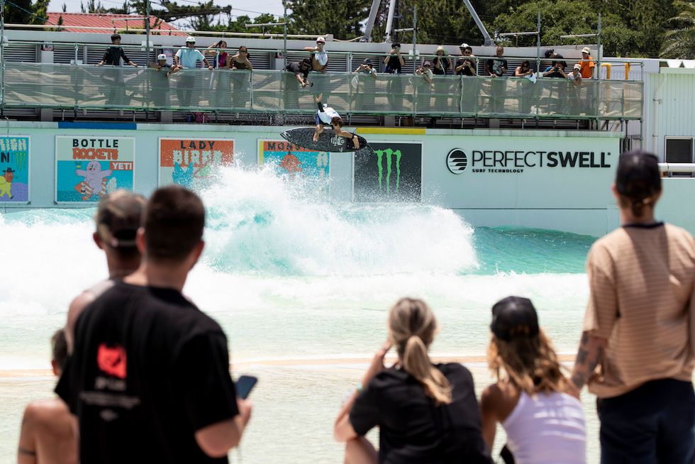Surfer catching air in wave pool, watched by onlookers, PerfectSwell sign in background.